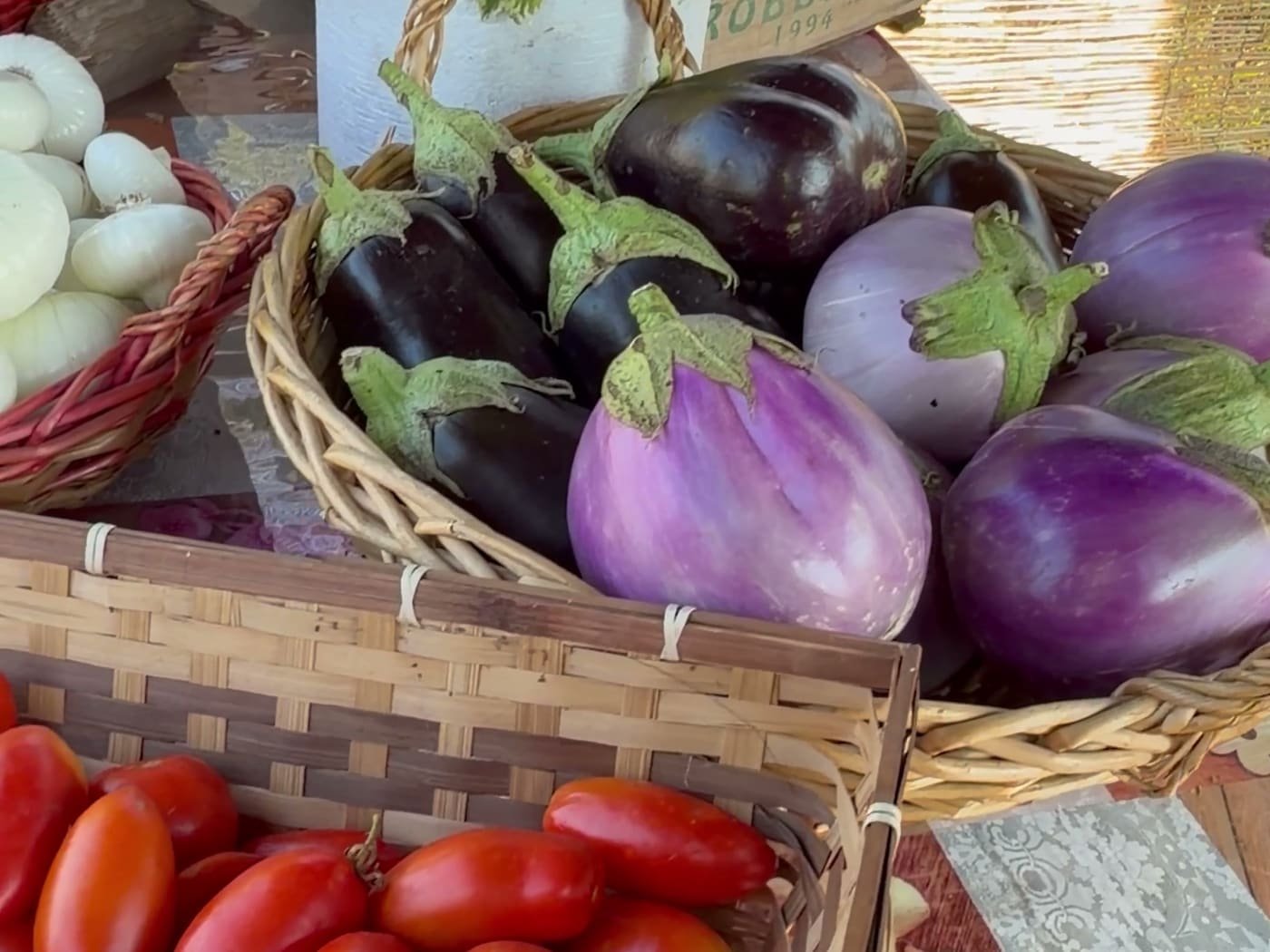 Ceste di melanzane scure e chiare accanto a una cassetta di pomodori perini e a un cesto di cipolle bianche